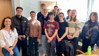 Eine Gruppe junger Menschen steht lächelnd und posiert für ein Foto in einem Raum. Einige tragen Brillen, Jacken und Turnschuhe. Die Person in der Mitte trägt ein Shirt mit Logo.