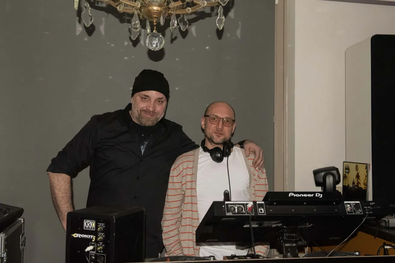 Zwei Männer stehen neben einer DJ-Anlage, einer trägt eine schwarze Mütze und der andere Kopfhörer, lächeln für ein Foto in einem Raum mit einer Kristallleuchte und einer grauen Wand.