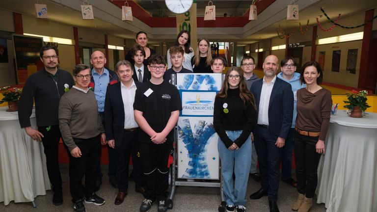 Eine Gruppe von Menschen, einschließlich Studenten und Fakultätsmitgliedern, steht für ein Foto zusammen, wobei zwei zentrale Figuren schwarze Shirts tragen und lächeln. Ein Banner mit blauen und weißen Designs steht hinter ihnen.