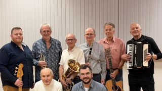 Eine Gruppe älterer Musiker, einige mit Brille, posiert für ein Foto mit verschiedenen Instrumenten wie Trompete, Saxophon und Gitarre.
