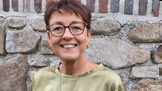 Eine Frau mit kurzen Haaren und Brille lächelt vor einer Steinmauer. Sie trägt ein grünes Shirt und eine Halskette mit Anhänger.