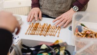 Eine Person ordnet kleine hölzerne Stricknadeln auf einem Tisch an, umgeben von Bastelmaterialien.