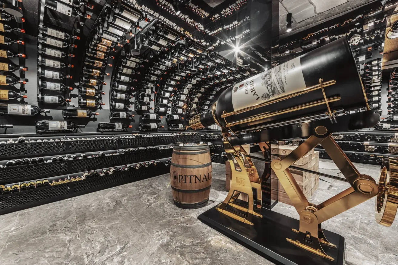 Ein Innenraumfoto eines großen Weinkellers, gefüllt mit zahlreichen Weinflaschen an den Regalen. Eine große Weinflasche mit dem Etikett Chateau Lafite ist prominent auf einem goldenen Ständer ausgestellt.