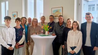 Eine Gruppe von Menschen steht um einen Tisch mit Blumen in einer Vase und lächelt für ein Foto. Der Tisch hat Gläser und eine grüne Tischdecke. Dahinter hängt ein gerahmtes Bild an der Wand.