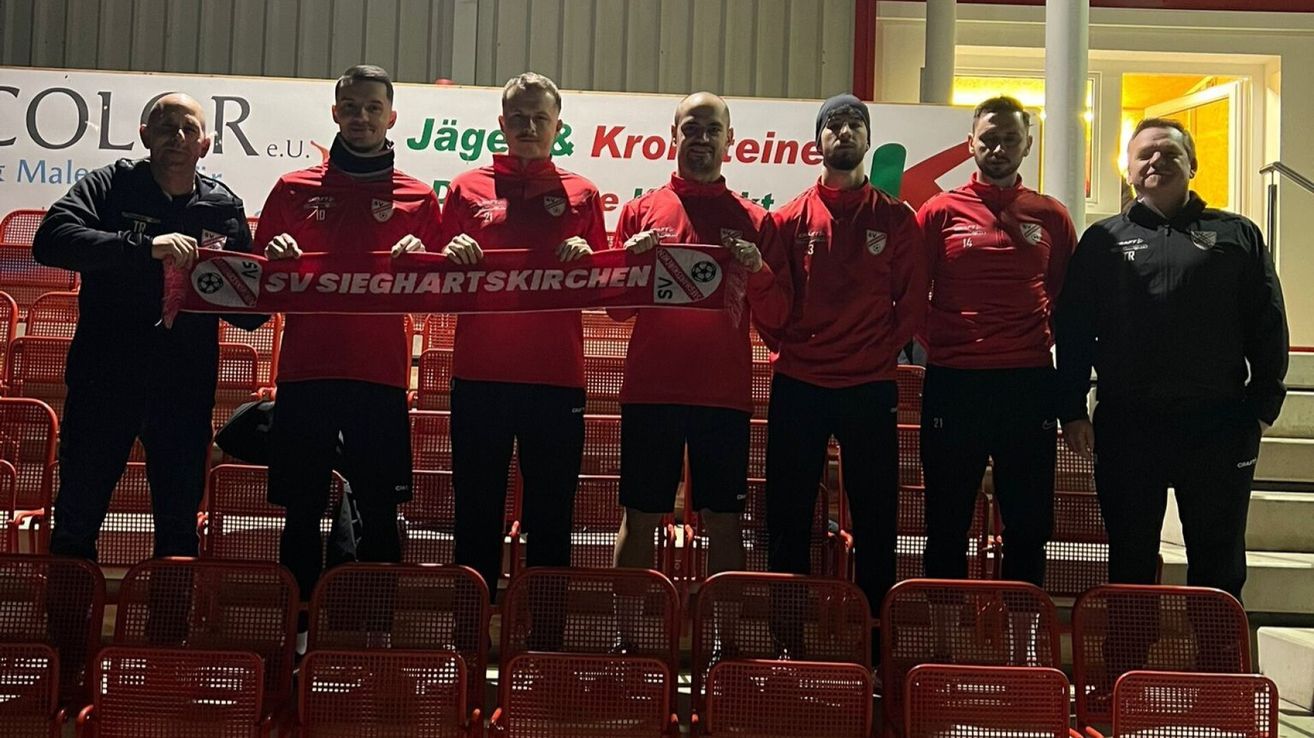 Fünf Männer in roten Uniformen stehen in einem Stadion und halten ein Banner mit der Aufschrift 'VS SIEGHARTSKIRCHEN'. Sie alle lächeln und posieren für ein Foto.
