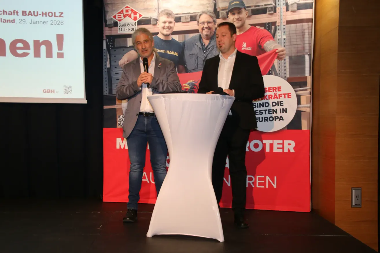 Zwei Männer stehen auf einer Bühne bei einer Veranstaltung. Einer spricht in ein Mikrofon an einem Podium. Ein großer Banner mit Text auf Deutsch steht hinter ihnen.