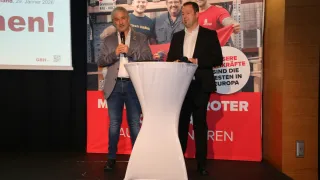 Zwei Männer stehen auf einer Bühne bei einer Veranstaltung. Einer spricht in ein Mikrofon an einem Podium. Ein großer Banner mit Text auf Deutsch steht hinter ihnen.