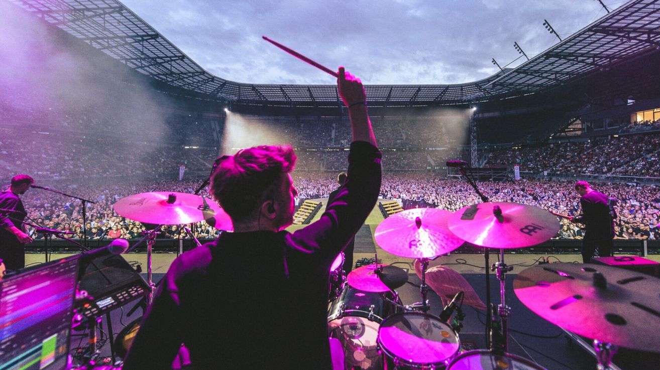Ein Schlagzeuger tritt bei einem großen Konzert mit einem farbenfrohen Bühnenaufbau auf, der von lila Lichtern beleuchtet wird, vor einem jubelnden Publikum.