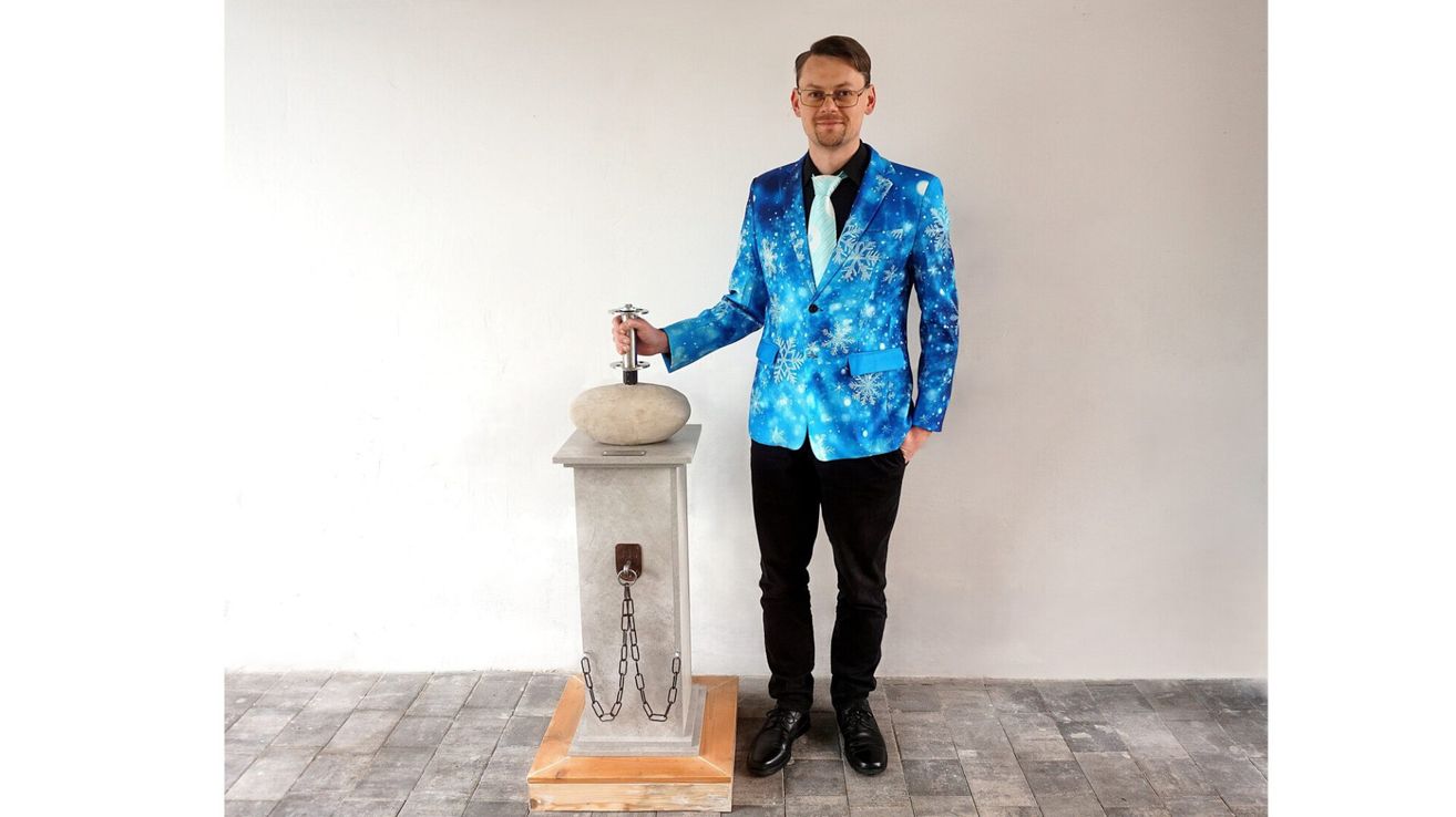 Ein Mann in einem blauen Anzug mit Schneeflockenmuster hält ein Metallobjekt mit einem Stein darauf. Er steht neben einem Betonsockel mit Ketten auf einem gefliesten Boden.