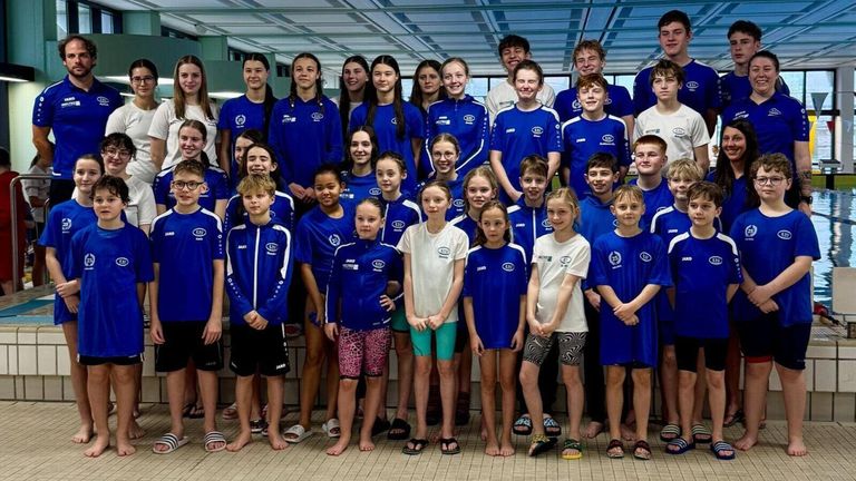 Eine Gruppe junger Schwimmer, sowohl Jungen als auch Mädchen, in blauen und weißen Uniformen, stehen zusammen auf der Poolbank und lächeln für ein Gruppenfoto.