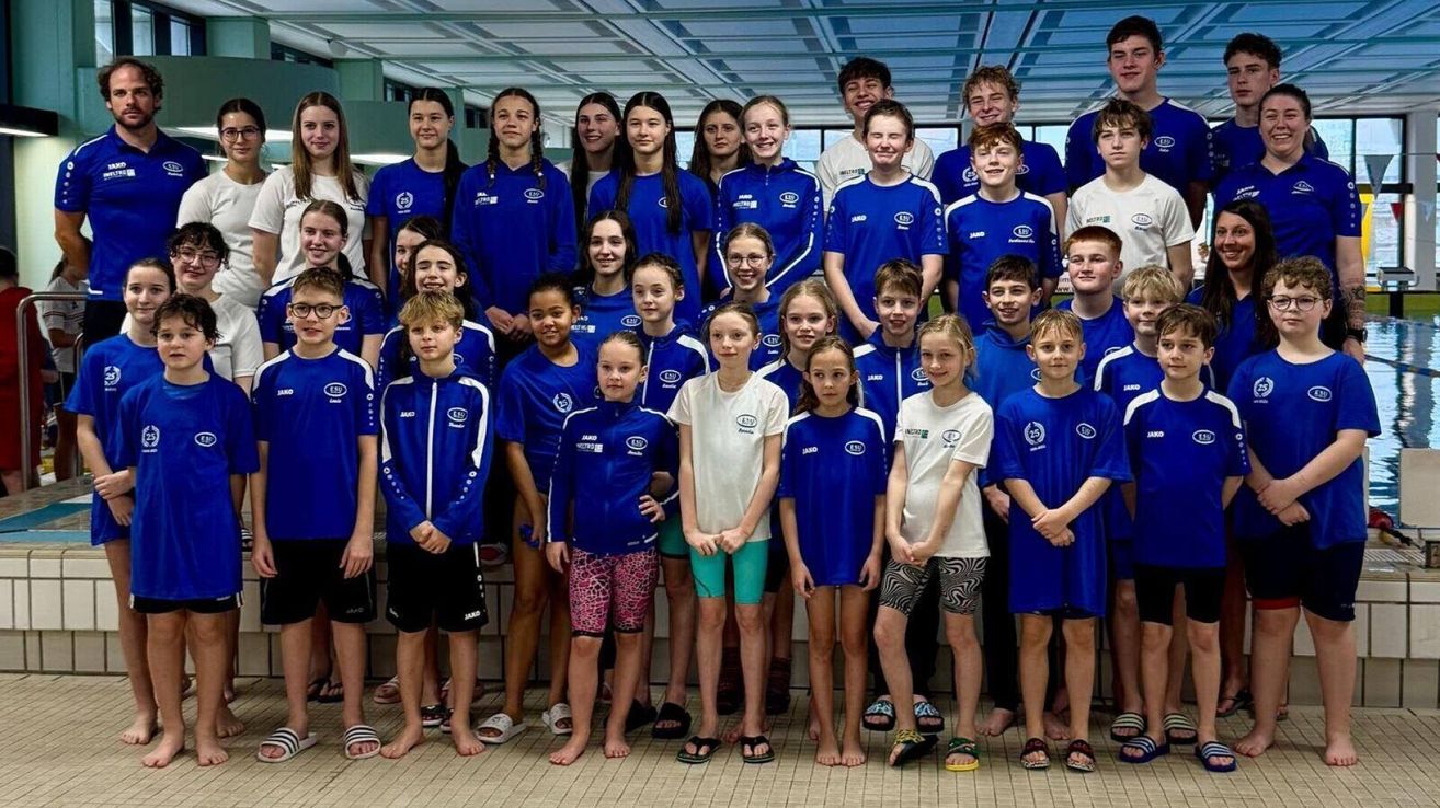 Eine Gruppe junger Schwimmer, sowohl Jungen als auch Mädchen, in blauen und weißen Uniformen, stehen zusammen auf der Poolbank und lächeln für ein Gruppenfoto.