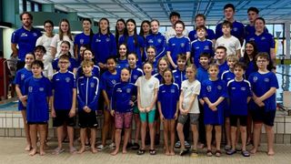 Eine Gruppe junger Schwimmer, sowohl Jungen als auch Mädchen, in blauen und weißen Uniformen, stehen zusammen auf der Poolbank und lächeln für ein Gruppenfoto.