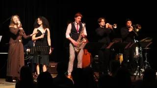 Eine Band tritt auf der Bühne auf. Eine Frau hält ein Mikrofon, während ein Mann Saxophon spielt. Ein anderer Mann spielt die Trompete. Dahinter sind ein Cello und Trommeln aufgebaut. Das Publikum schaut aus dem Dunkeln zu.