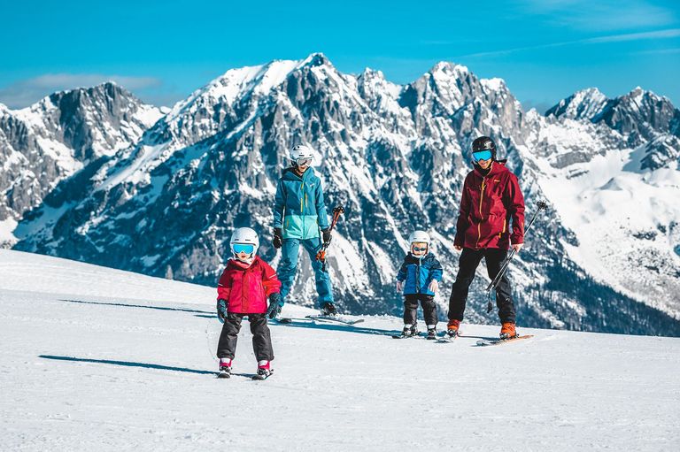 Eine vierköpfige Familie, zwei Erwachsene und zwei Kinder, skifahren auf einem verschneiten Berg mit blauem Himmel. Die Erwachsenen tragen Skiausrüstung, während die Kinder Helme und Schutzbrillen tragen.