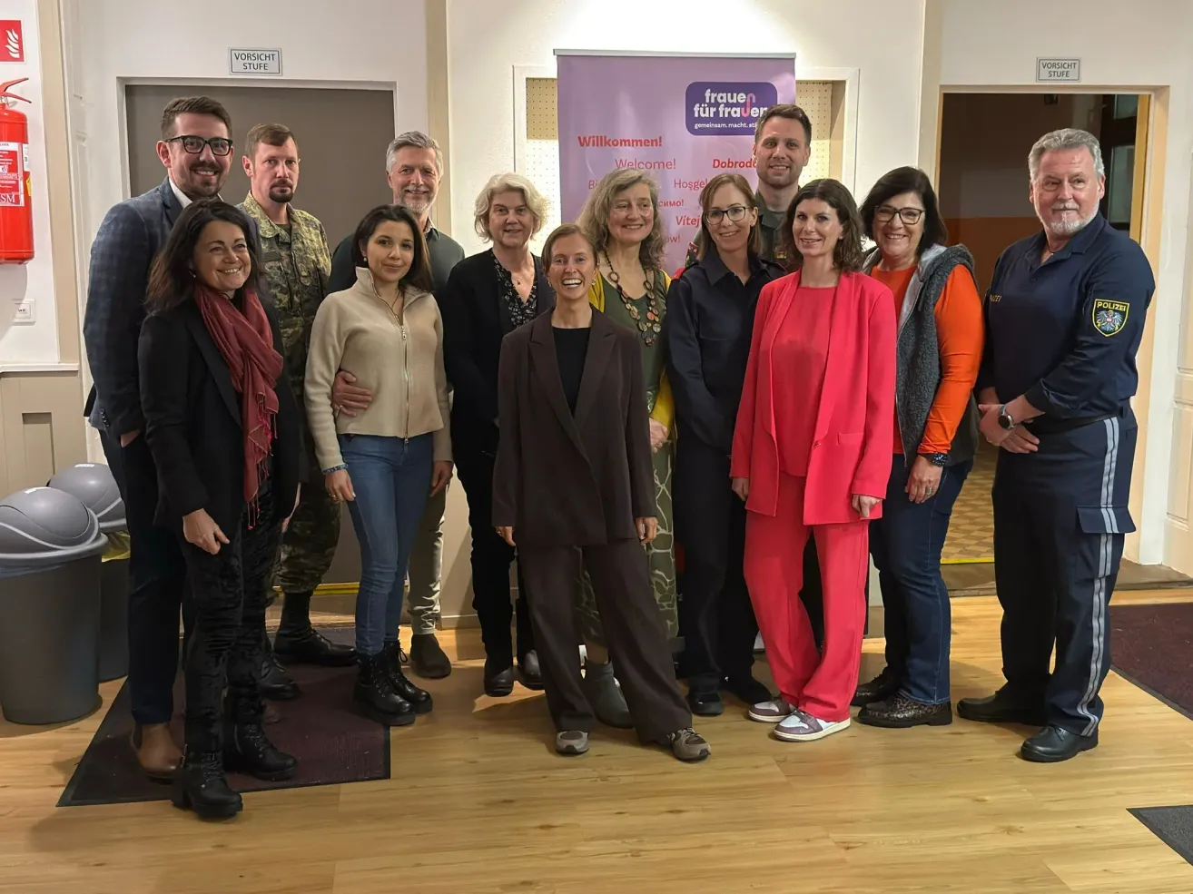 Eine Gruppe von Menschen steht für ein Foto zusammen, in verschiedenen Outfits, in einem Raum mit einem Banner dahinter.