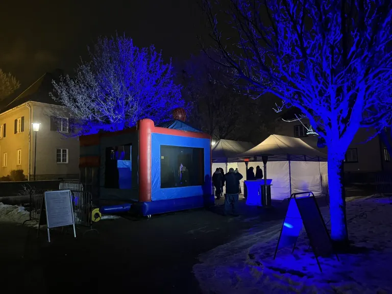 Ein blaues aufblasbares Zelt ist bei Nacht mit blauen Lichtern beleuchtet. In der Nähe stehen Menschen in der Nähe eines weißen Zeltes, und es gibt Bäume mit blauen Lichtern.