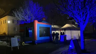 Ein blaues aufblasbares Zelt ist bei Nacht mit blauen Lichtern beleuchtet. In der Nähe stehen Menschen in der Nähe eines weißen Zeltes, und es gibt Bäume mit blauen Lichtern.