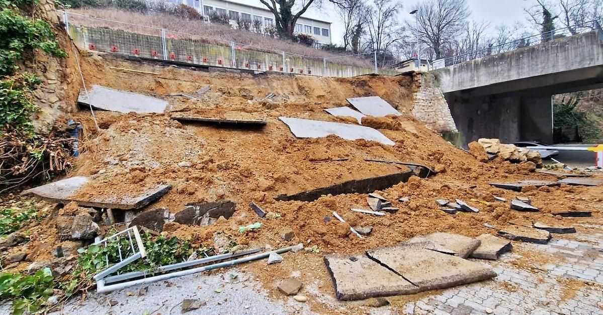 Ein Gelände mit einem eingestürzten Hang, verstreuten Trümmern und beschädigten Strukturen. Bäume und ein Gebäude sind im Hintergrund zu sehen.
