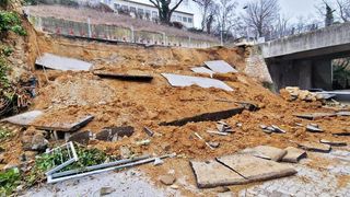 Ein Gelände mit einem eingestürzten Hang, verstreuten Trümmern und beschädigten Strukturen. Bäume und ein Gebäude sind im Hintergrund zu sehen.