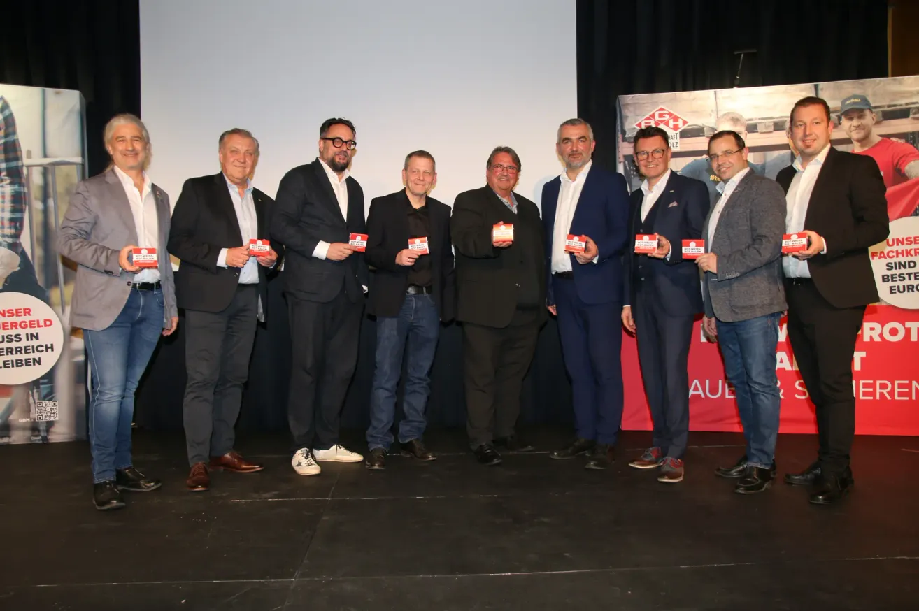 Eine Gruppe von Männern in Anzügen steht auf einer Bühne und hält kleine Karten in der Hand. Sie alle lächeln und scheinen für ein Foto zu posieren.