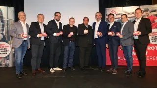 Eine Gruppe von Männern in Anzügen steht auf einer Bühne und hält kleine Karten in der Hand. Sie alle lächeln und scheinen für ein Foto zu posieren.