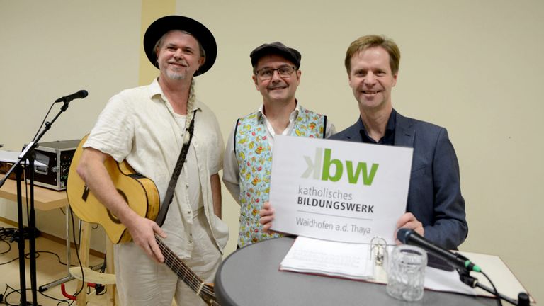 Drei Männer posieren für ein Foto; einer hält ein Schild für 'Katholisches Bildungswerk Waidhofen a.d. Thaya'. Ein Mann hält eine Gitarre, während die anderen lässig gekleidet sind.