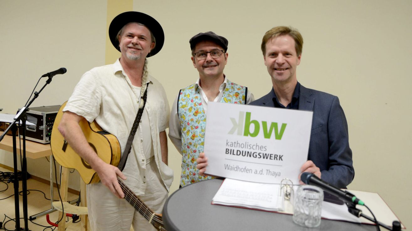 Drei Männer posieren für ein Foto; einer hält ein Schild für 'Katholisches Bildungswerk Waidhofen a.d. Thaya'. Ein Mann hält eine Gitarre, während die anderen lässig gekleidet sind.