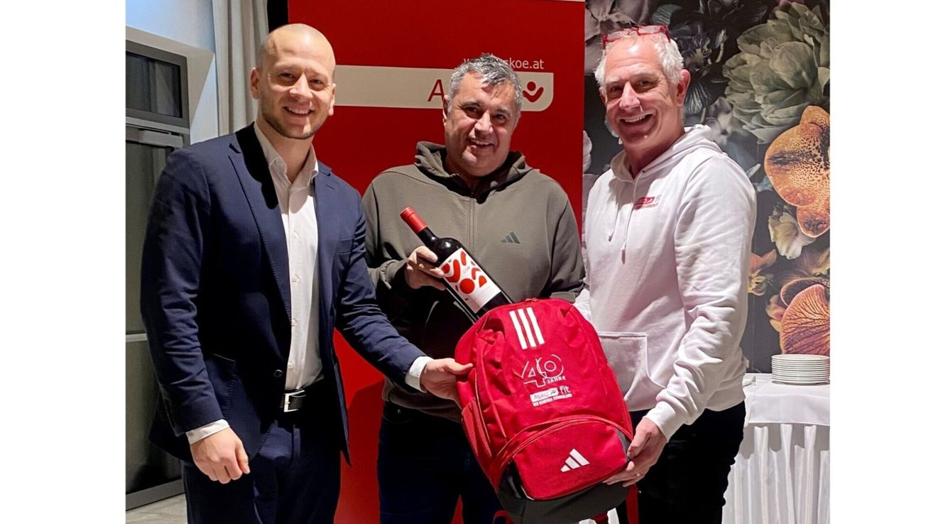 Drei Männer lächeln und posieren für ein Foto. Einer hält einen roten Rucksack und eine Flasche. Der Mann auf der linken Seite trägt einen blauen Anzug, und der Mann auf der rechten Seite trägt eine weiße Kapuzenjacke. Ein roter Banner ist hinter ihnen.