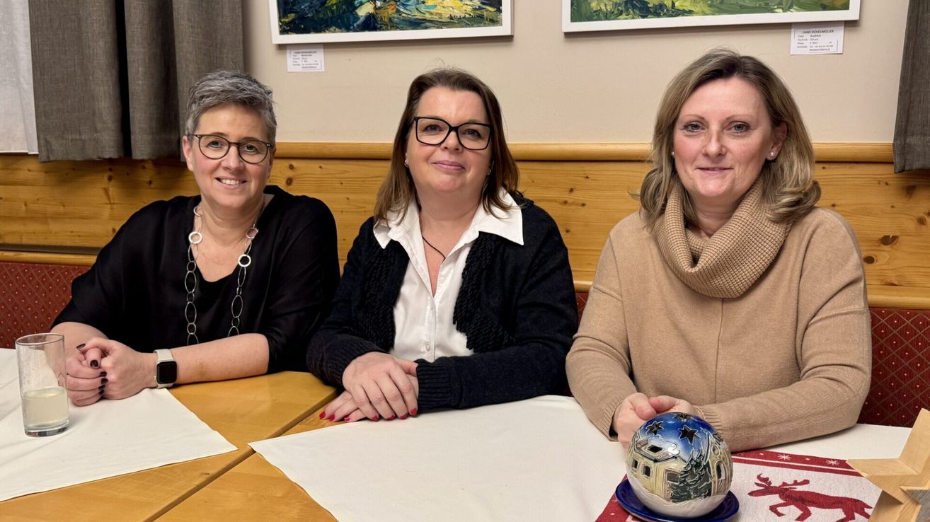 Drei Frauen sitzen lächelnd an einem Tisch. Eine hält eine Keramikkugel. Hinter ihnen hängen zwei gerahmte Gemälde an einer beigefarbenen Wand.