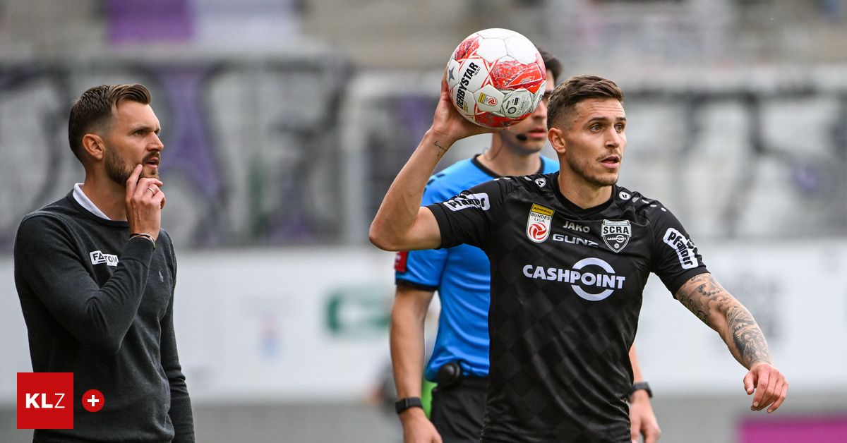 Ein Fußballspieler hält während eines Spiels einen Fußball hoch und trägt ein schwarzes Trikot mit dem Wort Cashpoint. Hinter ihm beobachtet ein weiterer Spieler in einem blauen Trikot.