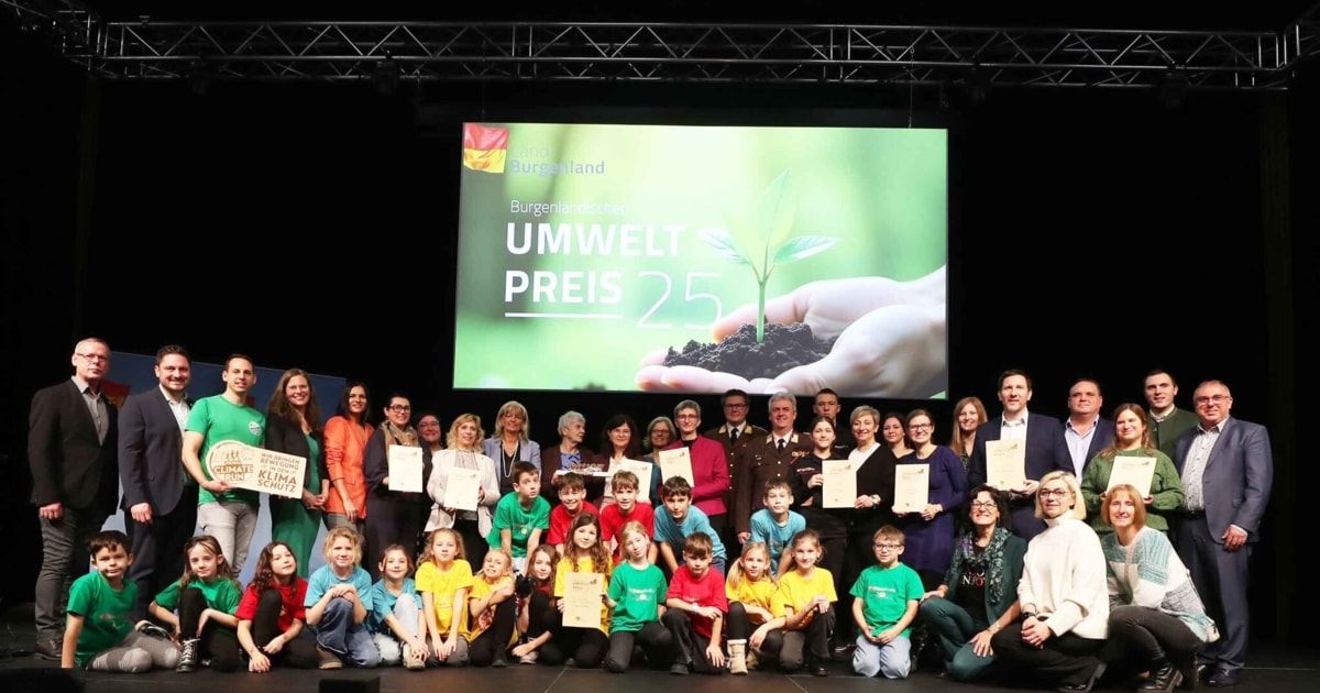 Eine Gruppe von Menschen, einschließlich Erwachsener und Kindern, posiert für ein Foto auf einer Bühne mit einer großen Leinwand, die eine Pflanze und die Worte 'Umwelt Preis 25' zeigt. Die Kinder halten Urkunden.