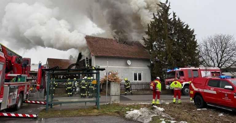 Ein Hausbrand mit Rauch, der aus dem Dach strömt. Feuerwehrleute stehen draußen mit Ausrüstung.