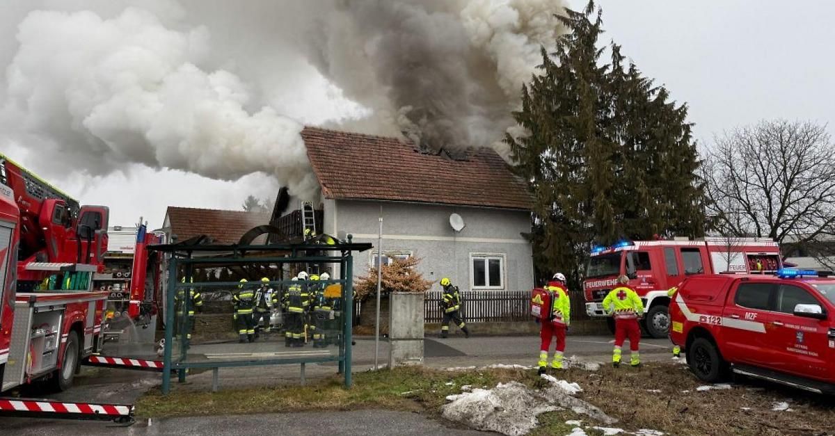 Ein Hausbrand mit Rauch, der aus dem Dach strömt. Feuerwehrleute stehen draußen mit Ausrüstung.