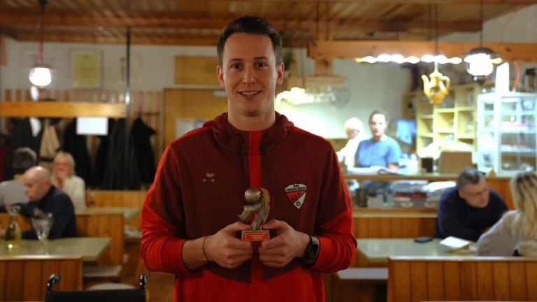 Ein Mann in einem roten Jackett mit einer Auszeichnung steht lächelnd in einem Raum mit Holzbalken und trägt eine Uhr. Im Hintergrund sind zwei Personen sichtbar, eine sitzt und die andere steht.