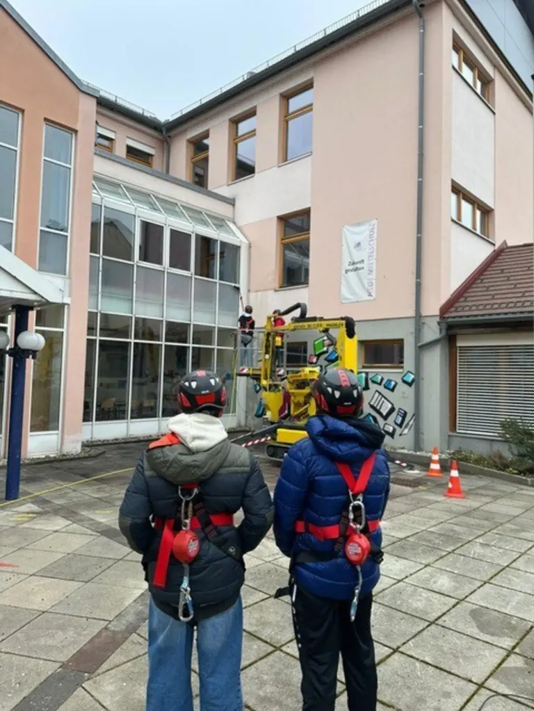 Zwei Personen in Sicherheitsausrüstung beobachten eine Person, die die Glasfenster eines Gebäudes mit einer Maschine reinigt.