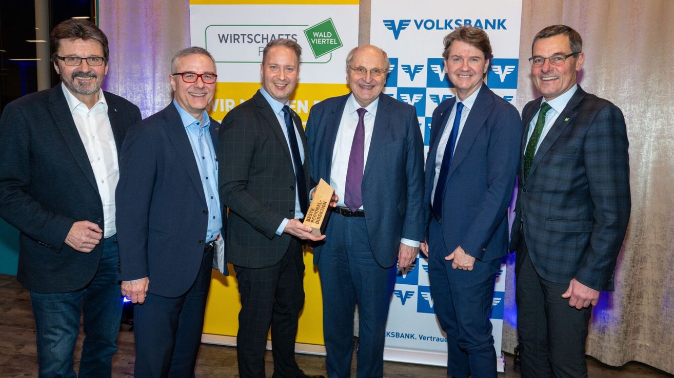 Fünf Männer in formeller Kleidung stehen vor einer Kulisse. Ein Mann hält eine Trophäe.