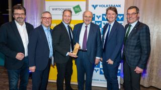 Fünf Männer in formeller Kleidung stehen vor einer Kulisse. Ein Mann hält eine Trophäe.