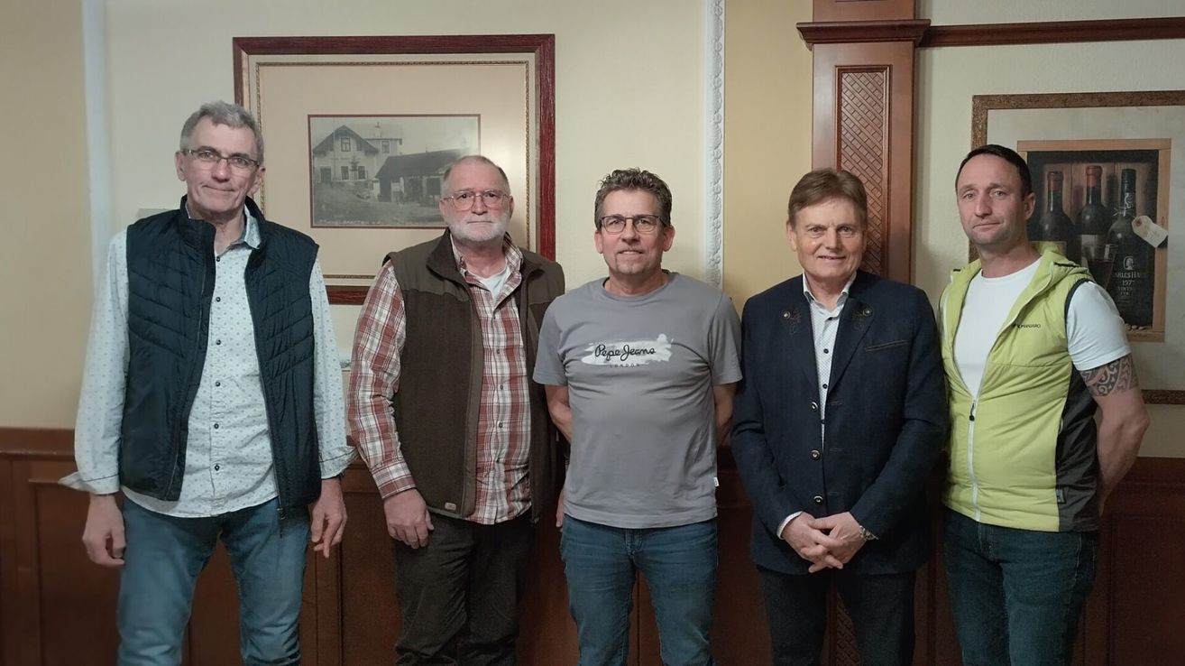 Vier Männer stehen vor einer Wand. Der Mann ganz links trägt eine braune Weste. Der zweite Mann hat eine Brille und ein kariertes Hemd. Der dritte Mann trägt ein graues T-Shirt. Der Mann ganz rechts trägt einen dunklen Blazer.