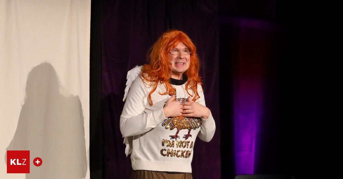 Eine Person mit roten Haaren, die ein weißes Shirt mit einem Hühnerdesign und Engelsflügeln trägt, lächelt auf der Bühne.