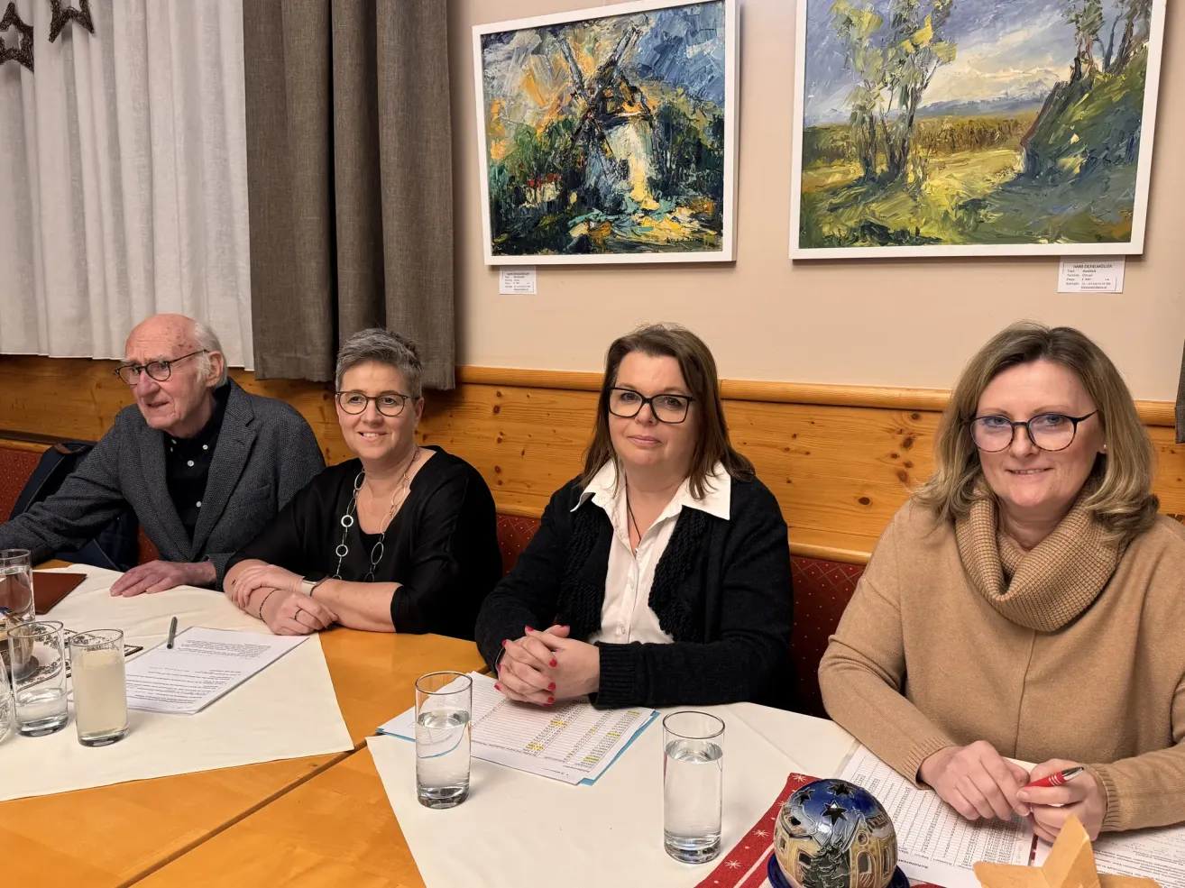 Vier Personen sitzen um einen Tisch herum, möglicherweise bei einer Besprechung. Papiere und Gläser stehen auf dem Tisch. Hinter ihnen hängen zwei Gemälde an der Wand.