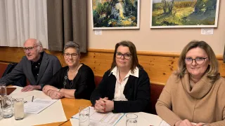 Vier Personen sitzen um einen Tisch herum, möglicherweise bei einer Besprechung. Papiere und Gläser stehen auf dem Tisch. Hinter ihnen hängen zwei Gemälde an der Wand.