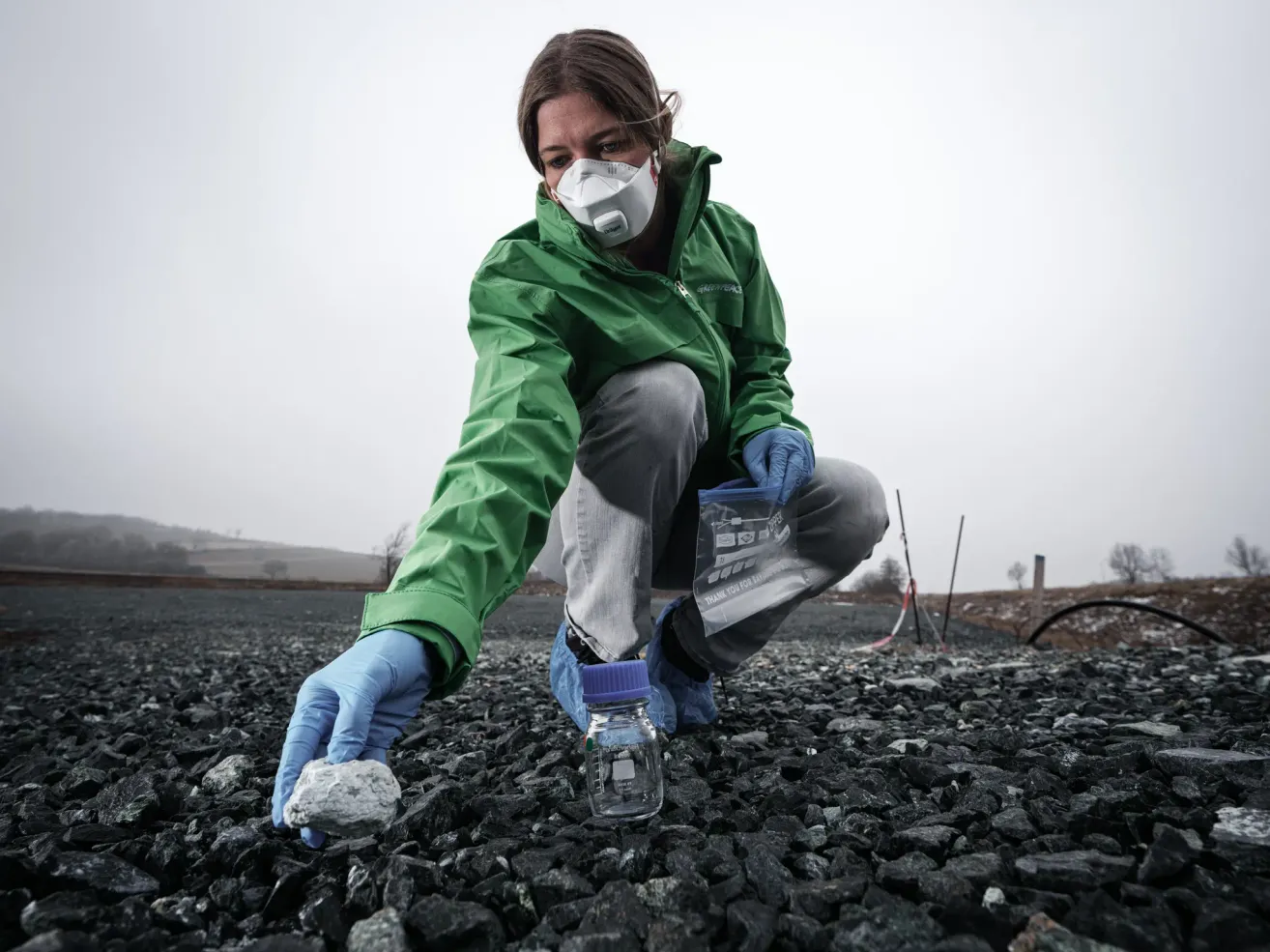 Eine Frau in einer grünen Jacke und einer weißen Maske kniet auf dem Boden, hält einen Felsen und einen Glasbehälter. Sie ist möglicherweise eine Wissenschaftlerin, die ein Experiment durchführt.