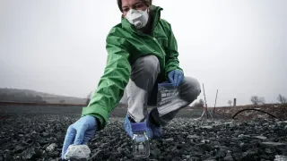 Eine Frau in einer grünen Jacke und einer weißen Maske kniet auf dem Boden, hält einen Felsen und einen Glasbehälter. Sie ist möglicherweise eine Wissenschaftlerin, die ein Experiment durchführt.
