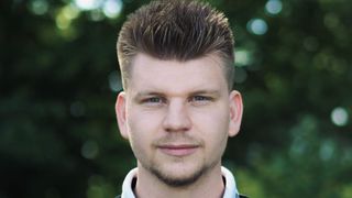 Ein Mann mit einem ordentlich gestylten Haarschnitt und Bart lächelt direkt in die Kamera, vor einem unscharfen grünen Hintergrund.