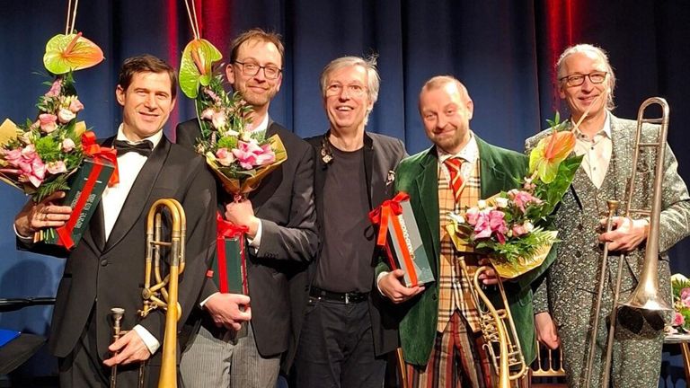 Fünf Männer in formeller Kleidung stehen lächelnd zusammen auf einer Bühne. Sie halten Geschenke und Blumen. Einer hält eine Posaune, und ein anderer einen Strauß aus rosa und grünen Blumen. Ein blauer Vorhang ist hinter ihnen.