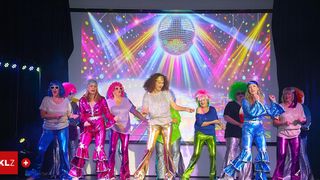Eine Gruppe von Frauen in Disco-Kostümen der 70er Jahre tritt auf der Bühne auf, mit einer leuchtenden Discokugel im Hintergrund.