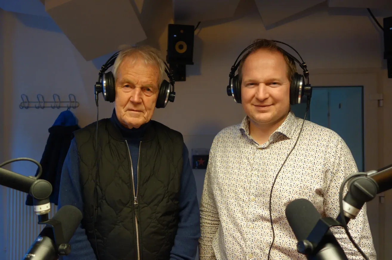 Zwei Männer stehen in einem Studio und tragen Kopfhörer. Einer ist älter mit einer schwarzen Weste, der andere ist jünger mit einem gepunkteten Hemd. Beide haben Mikrofone vor sich.