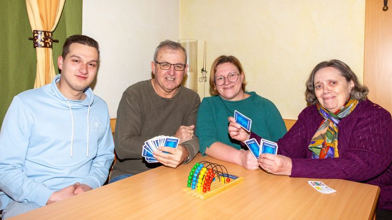 Vier Menschen sitzen um einen Holztisch, lächeln und halten Karten. Auf dem Tisch in der Mitte befindet sich ein Holzperlen-Labyrinth. Der Raum hat eine gelbe Wand und einen Kerzenständer.