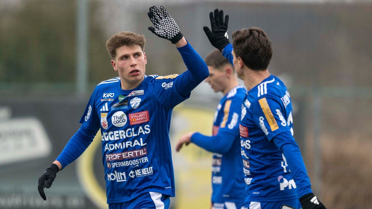 Ein Fußballspieler feiert und trägt ein blaues Trikot mit verschiedenen Werbeanzeigen. Zwei Teamkollegen sind in der Nähe, einer im Hintergrund, alle in blauen Uniformen.