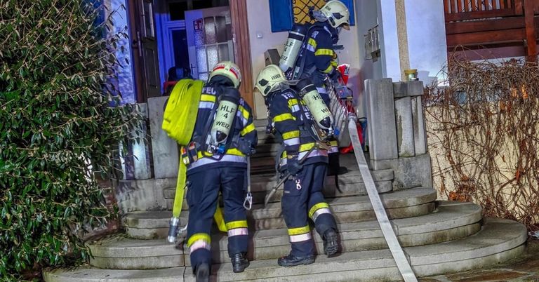 Drei Feuerwehrleute, die Helme tragen und Ausrüstung mit sich führen, erklimmen die Treppe, um ein Gebäude zu betreten. Sie haben Sauerstoffflaschen und einen Feuerlöscher bei sich.